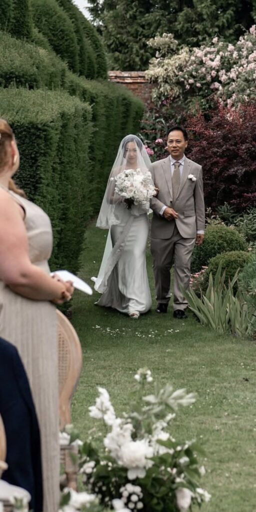 Minimalist Riki Dalal bride walking with dad through formal gardens in a sculpted satin wedding dress following couture bridal alterations and veil finishing