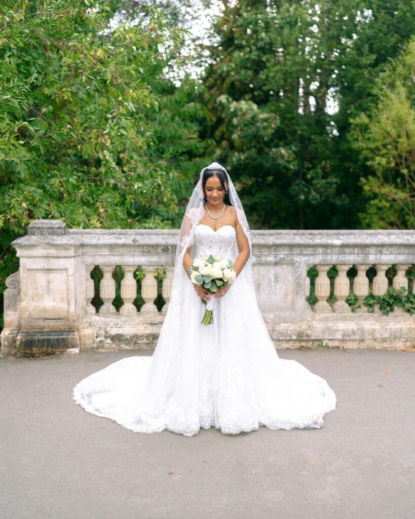 Bride wearing an Anna Sorrano Mercia lace wedding dress and holding a white rose bouquet beside a stone balustrade