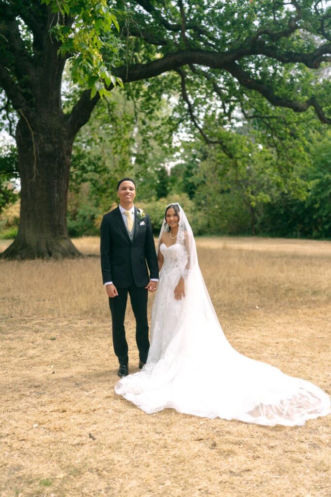Bride in an Anna Sorrano Mercia wedding dress holding hands with her groom beneath a tree, with her veil spread across the grass