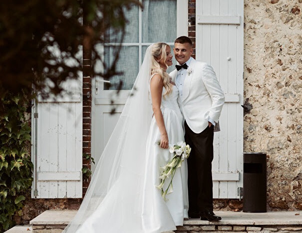 Full-length wedding portrait of the bride and groom beside white shutters