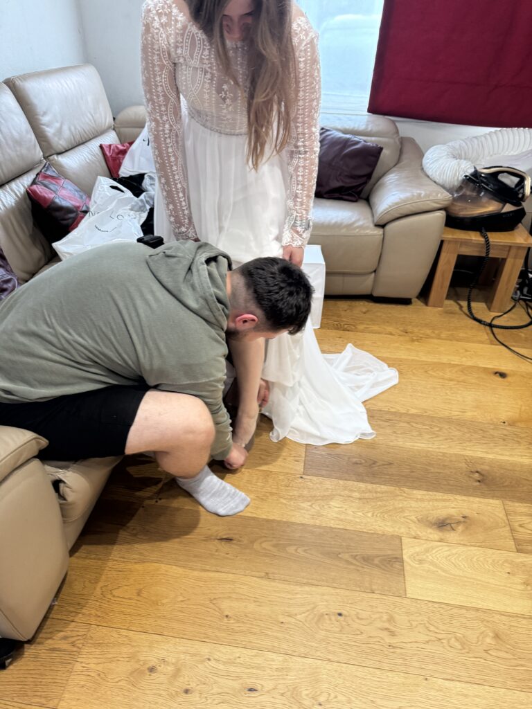 Groom helping the bride put on her wedding shoes during her bridal alterations fitting in Sutton