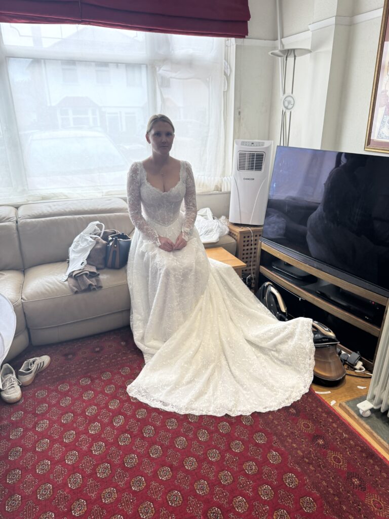 Intricately beaded lace wedding dress final try on of a Yedyna Estia bridal gown bride is sitting down at fitting day.